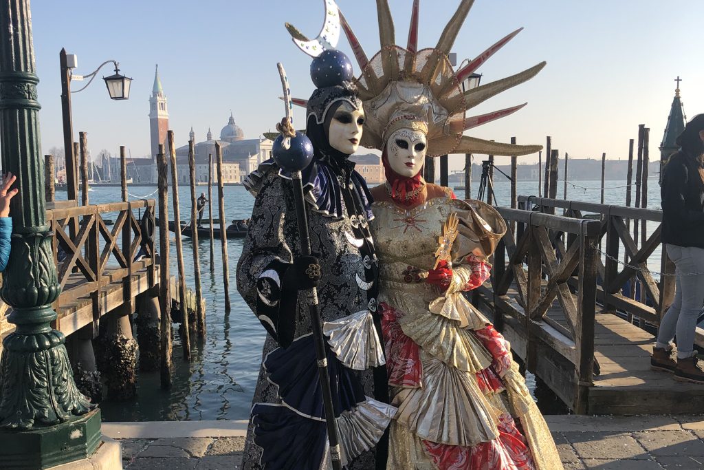 Two people in elaborate Venetian carnival costumes with ornate masks, feathers, and baroque-style embroidery, standing against a light-colored building wall during sunset
