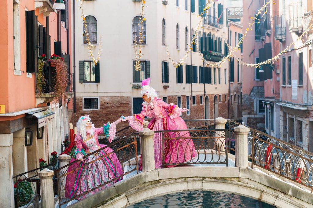 Two people in elaborate 18th-century-style pink costumes and white wigs posing on a small arched bridge over a canal in Venice, surrounded by historic buildings and festive string lights.