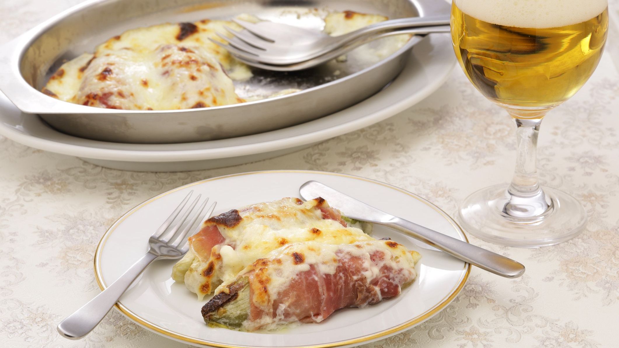 A white dish with two endives wrapped in ham and topped with melted cheese. The baking pan with the rest of the baked food is in the background.