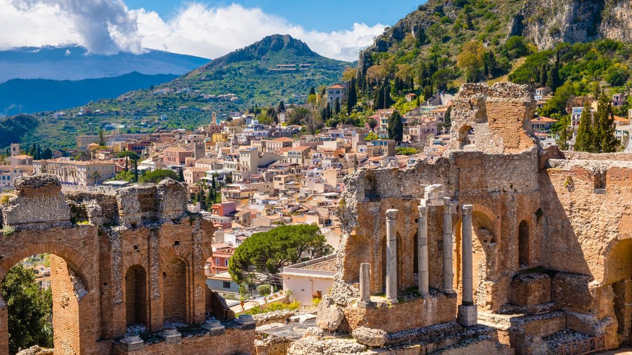 The ruins of an Ancient Greek theater, with the Old Town and Mount Etna covered with clouds in the background.