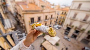A hand holds a traditional Sicilian cannolo over a balcony with a view of old Palermo rooftops.