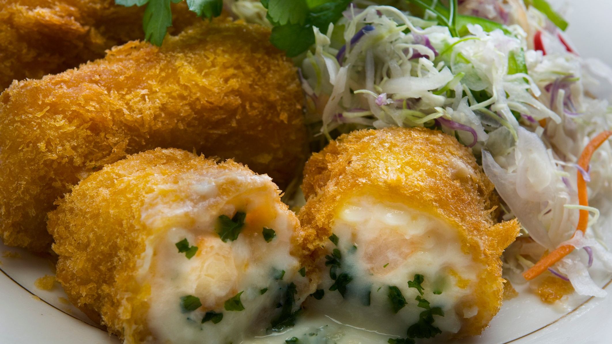 A close-up of a golden croquette cut in half. There's white sauce, shrimp, and herbs inside.