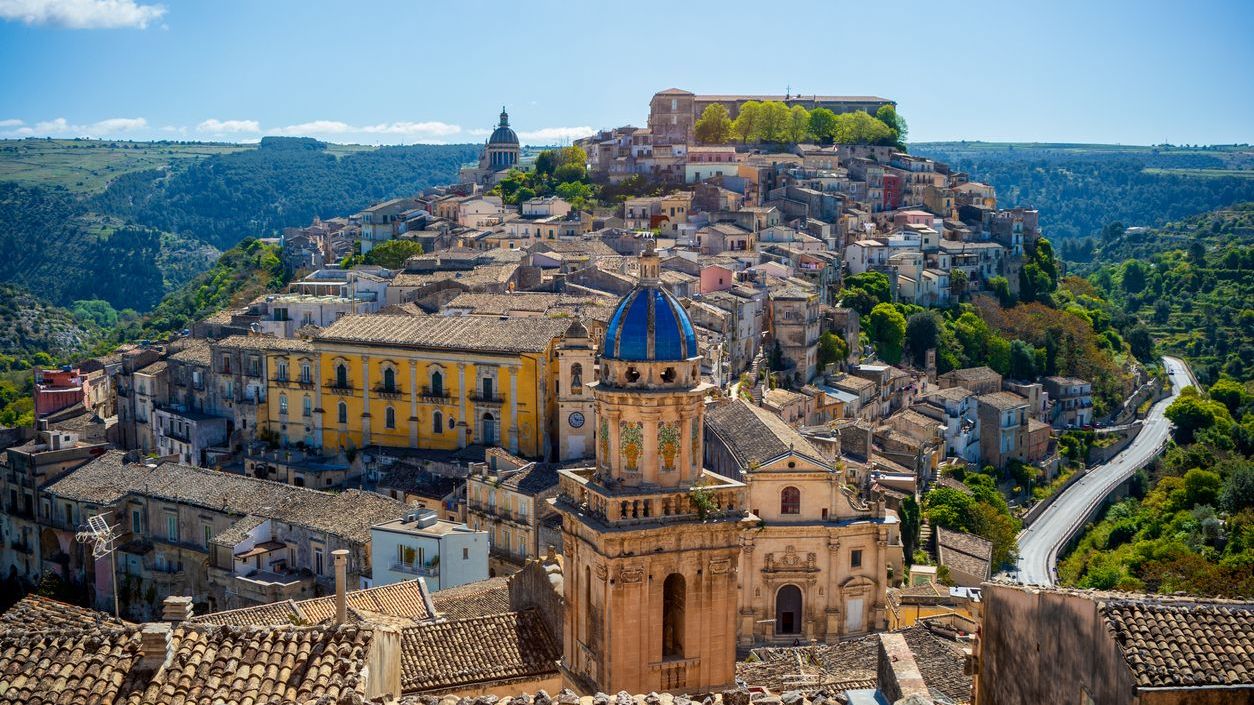 A panoramic view of the historic center of Ragusa, Sicily, Italy, with the tower of the blue-domed Santa Maria dell'Itria church, a UNESCO World Heritage Site, in the foreground. 