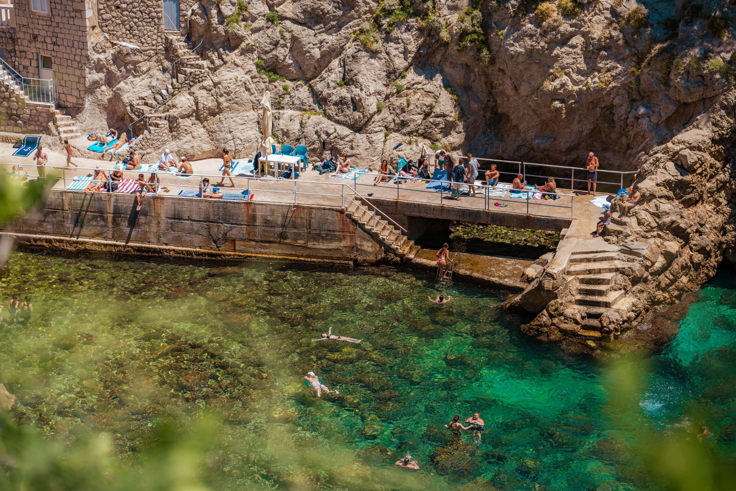 People sitting on the rocks and others swimming in emerald waters. 