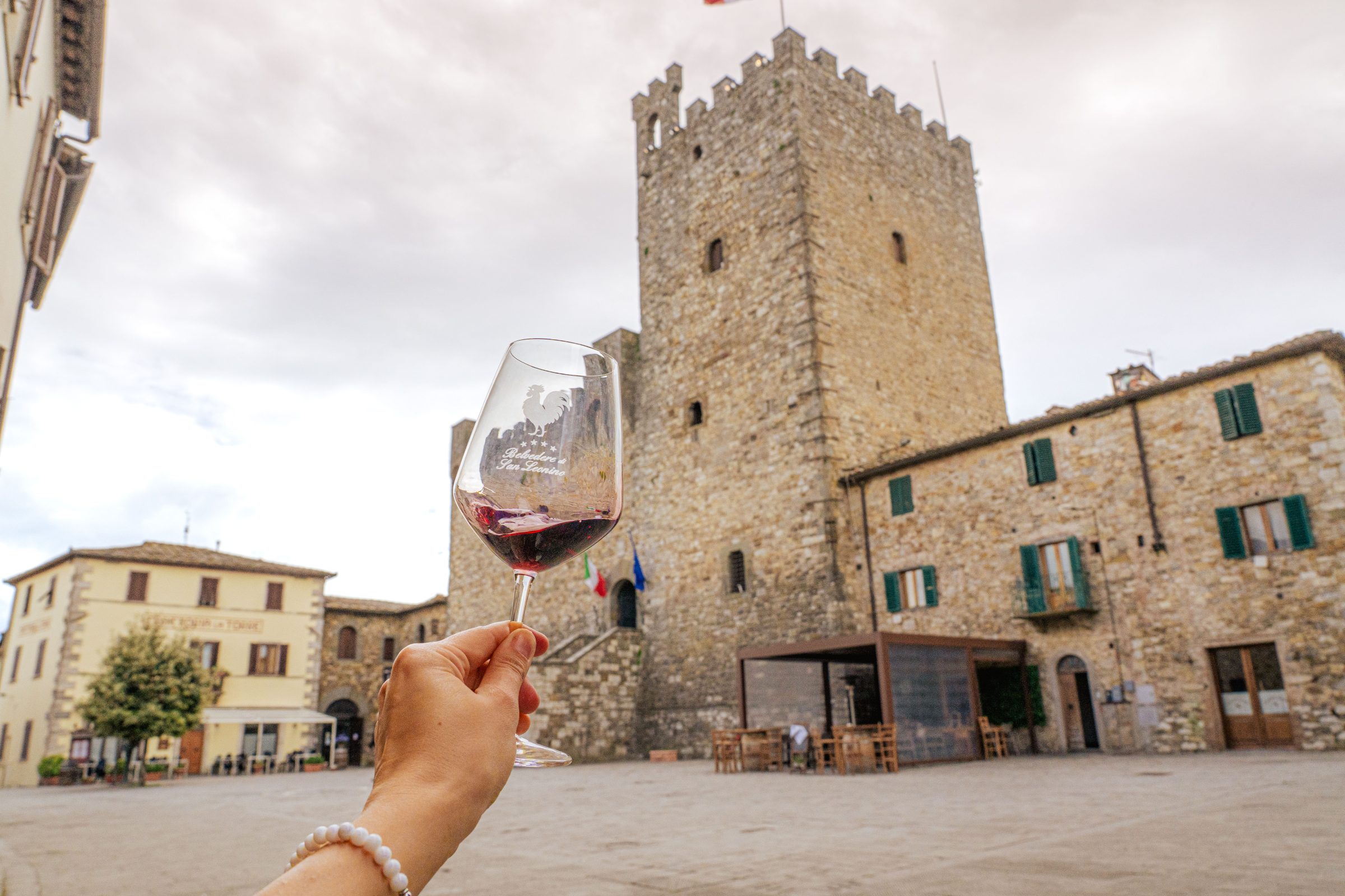 A hand is holding a glass of Chianti wine in front of a traditional Tuscan building.
