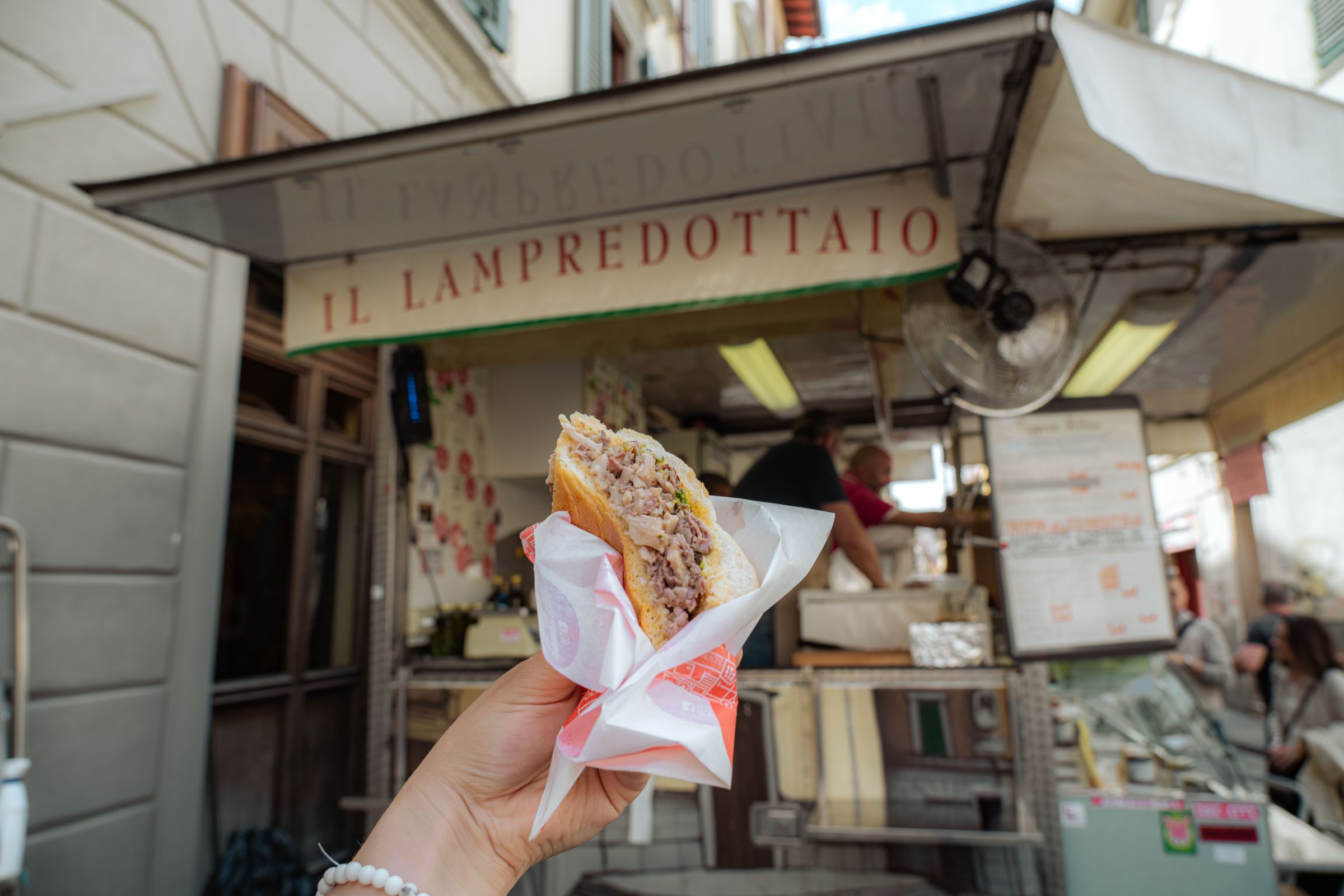 A hand is holding a lampredotto sandwich in front of a lampredotto food truck.