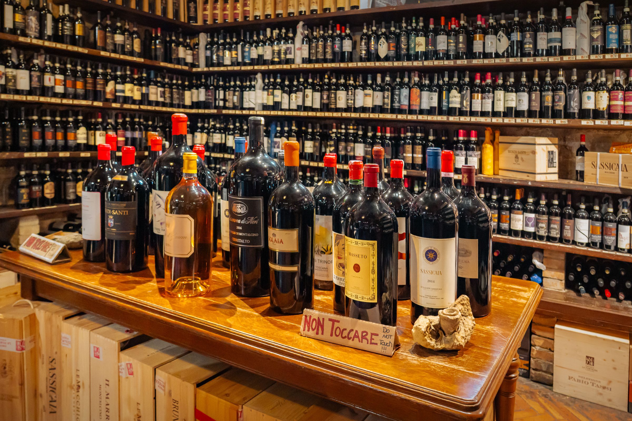A wooden table with many bottles of wine on it and shelves filled with wine bottles in the background.