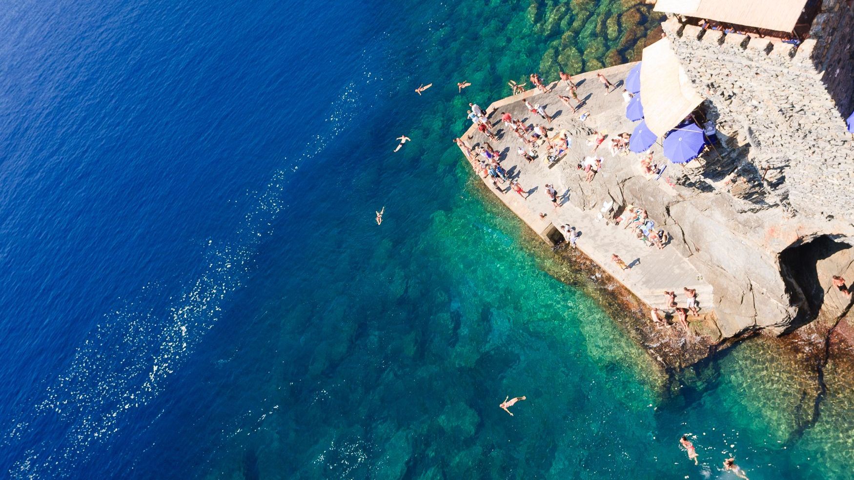 An aerial view of a rocky jetty from which people dive into the dark blue sea in Vernazza, one of the best places to visit in Cinque Terre. 