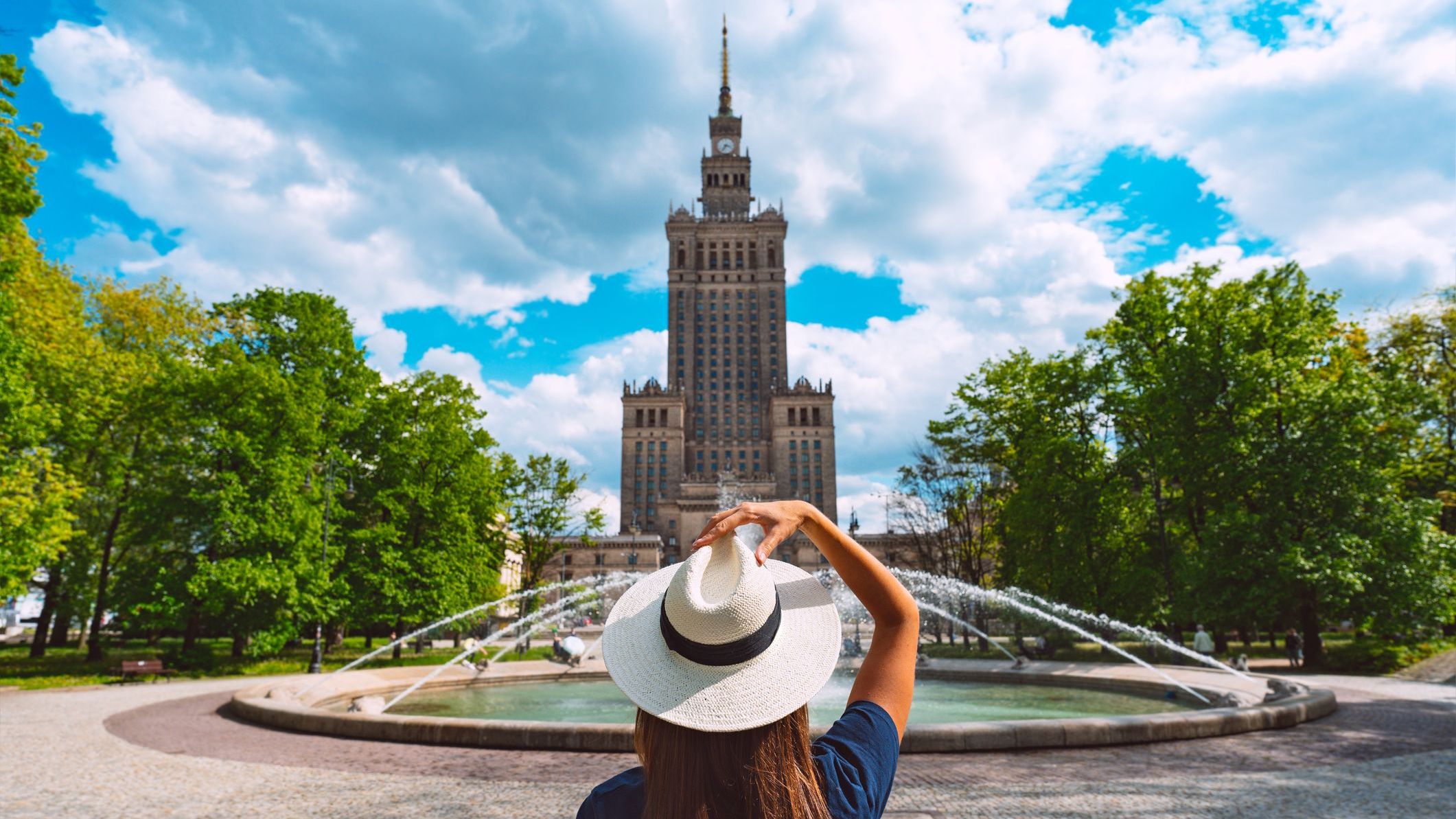 A young woman wearing a white sun hat is standing in front of the Palace of Culture and Science in Warsaw.