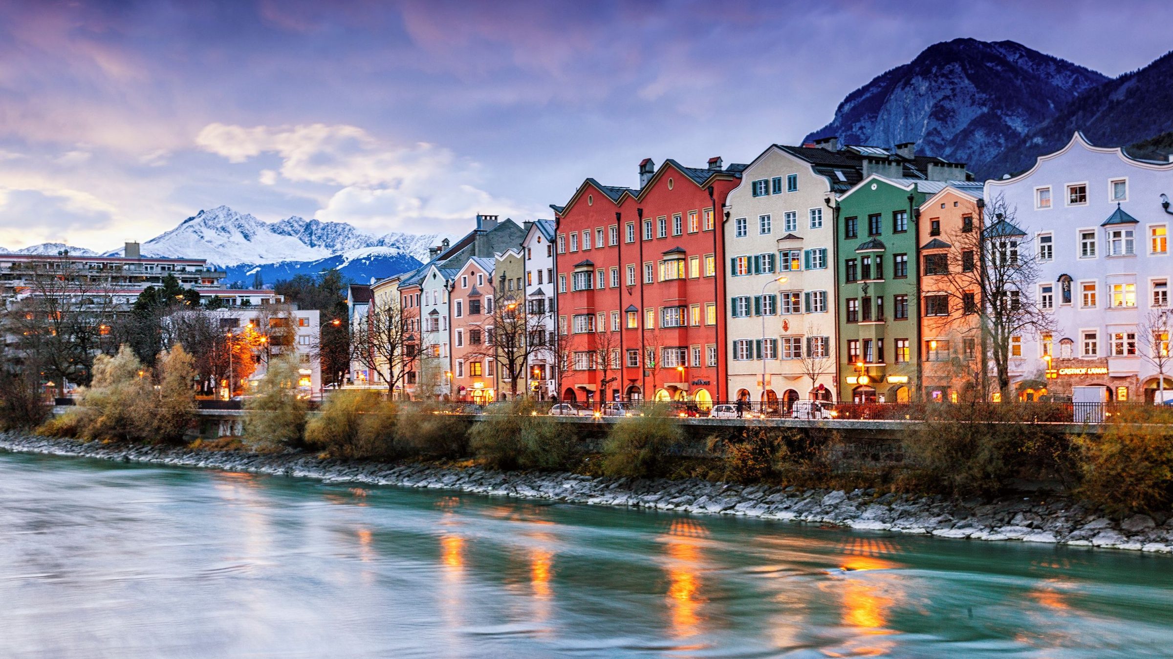A row of pastel-colored buildings along a river with snow-capped mountains in the background. 