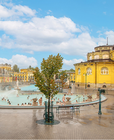 A photo of bathers enjoying an outdoor thermal pool surrounded by the distinct yellow buildings of the Neo-Baroque style Széchenyi Thermal Bath, the largest medicinal bath in Europe, which is supplied by two thermal springs on a sunny, summer day in Budapest, Hungary.