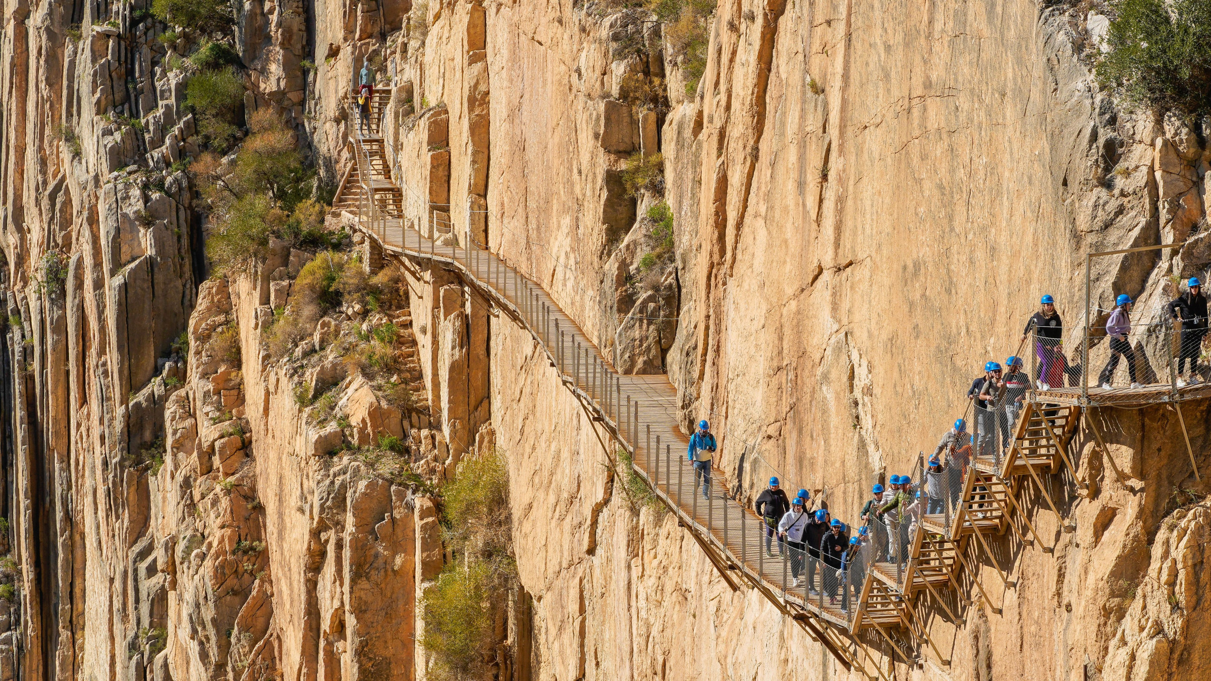 People wearing blue helmets are walking along a narrow wooden boardwalk pinned to a vertical limestone cliff.