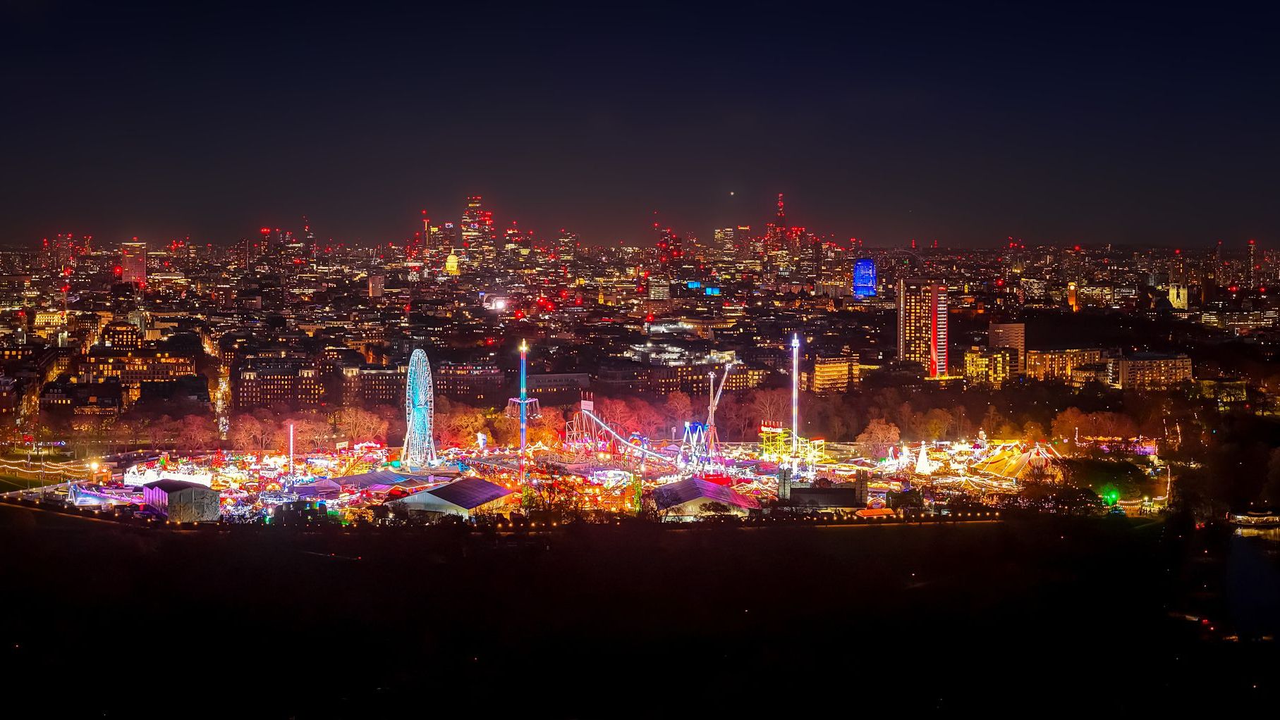 Vibrant Christmas fair lights glow in Hyde Park beneath the London skyline at dusk, featuring rides, a Ferris wheel, and festive cheer.