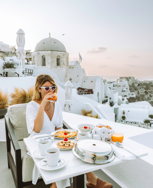 Woman enjoying a drink in Oia with whitewashed buildings and Aegean Sea views in the background.