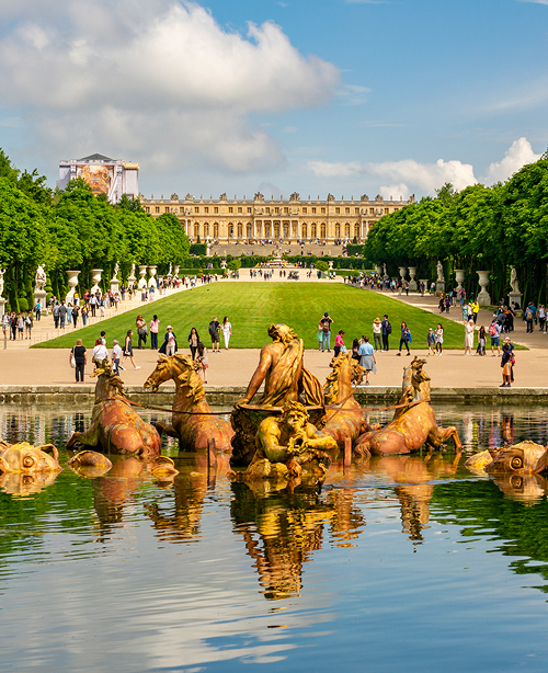 Expansive formal gardens at the Palace of Versailles in Versailles, featuring geometric hedges, fountains, and tree-lined pathways under a clear sky