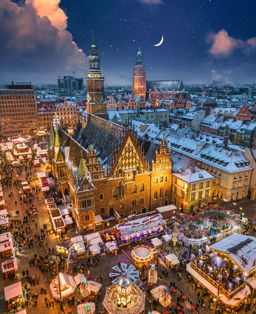 Festive European Christmas market with illuminated wooden stalls, holiday decorations, and crowds browsing seasonal gifts at night.