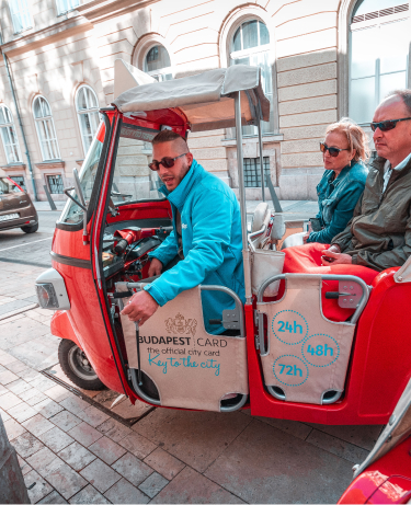 A guide driving two tourists through the streets of Budapest in a red tuk-tuk displaying the Budapest Card