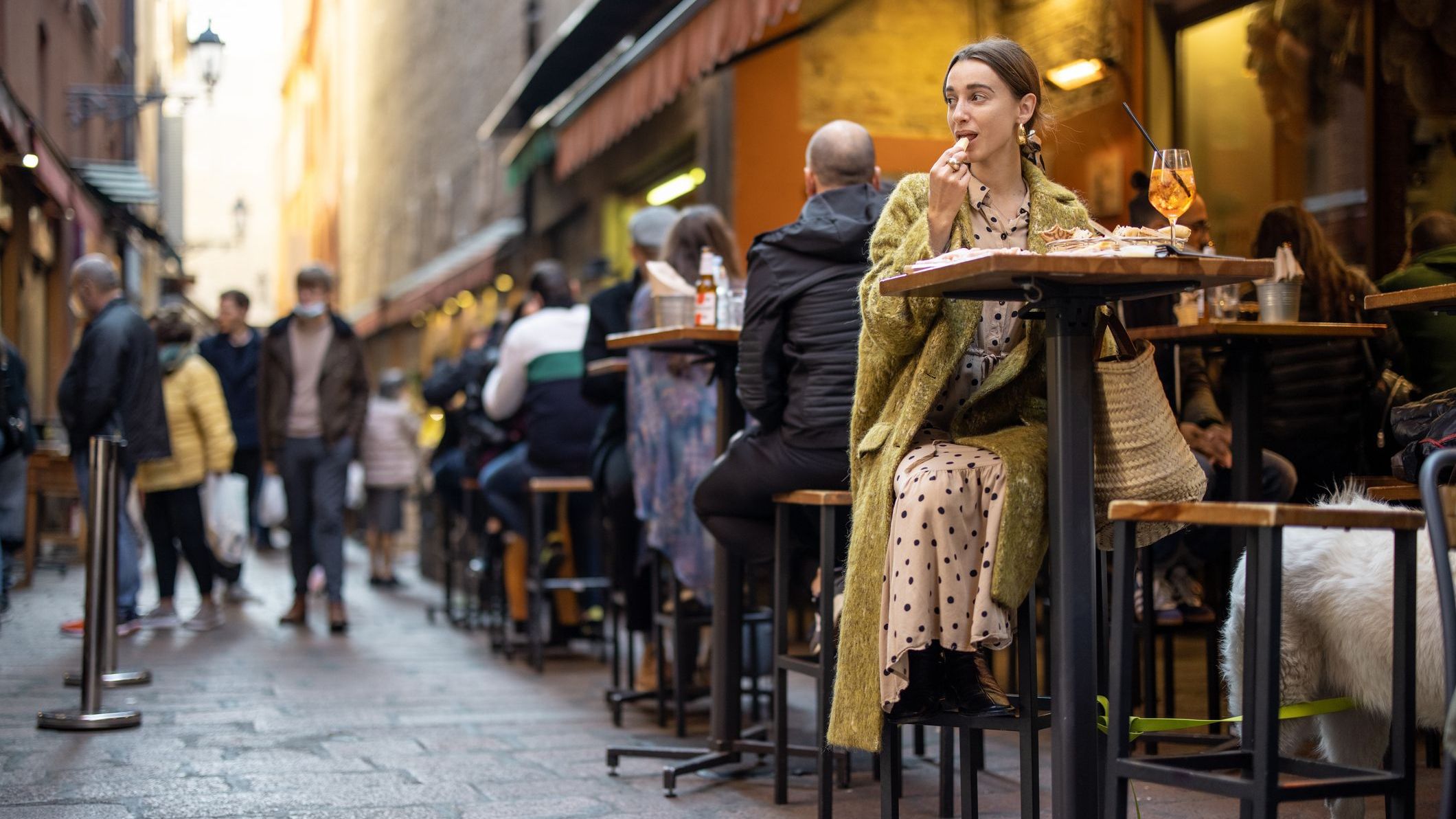 A woman sitting at an outdoor bar along a crowded street, eating nibbles and drinking an Aperol Spritz. 