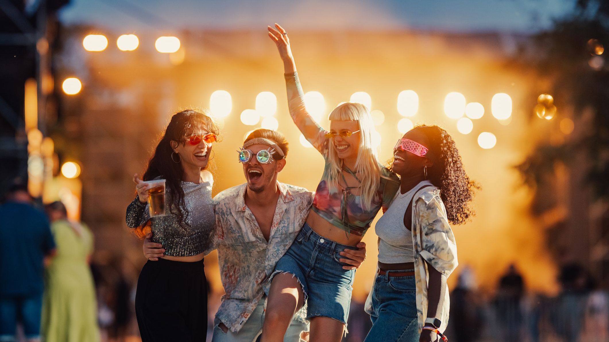 Four friends are laughing and having fun at an outdoor music festival, enjoying a live concert.