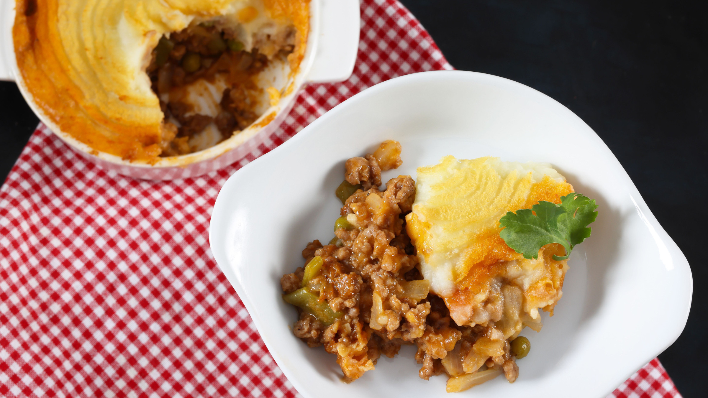 A pot of Shepherd's Pie and a piece of Shepherd's Pie served in a white plate on a red and white checkered tablecloth. 