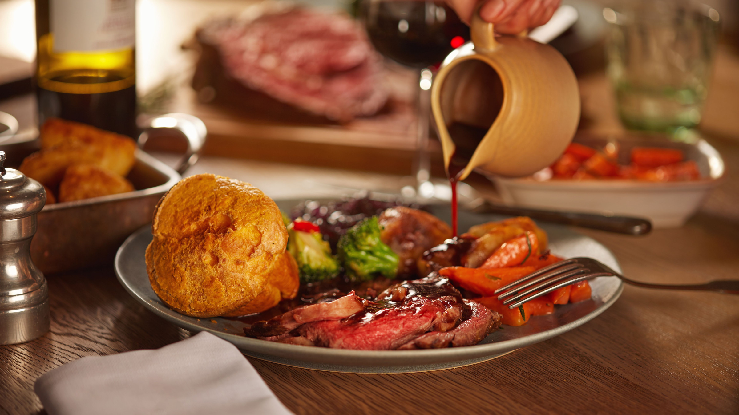 A hand is pouring gravy out of a small pot over a dish that contains roast meat, vegetables, and a Yorkshire pudding, one of the best things to try if you're wondering what to eat in the UK. 