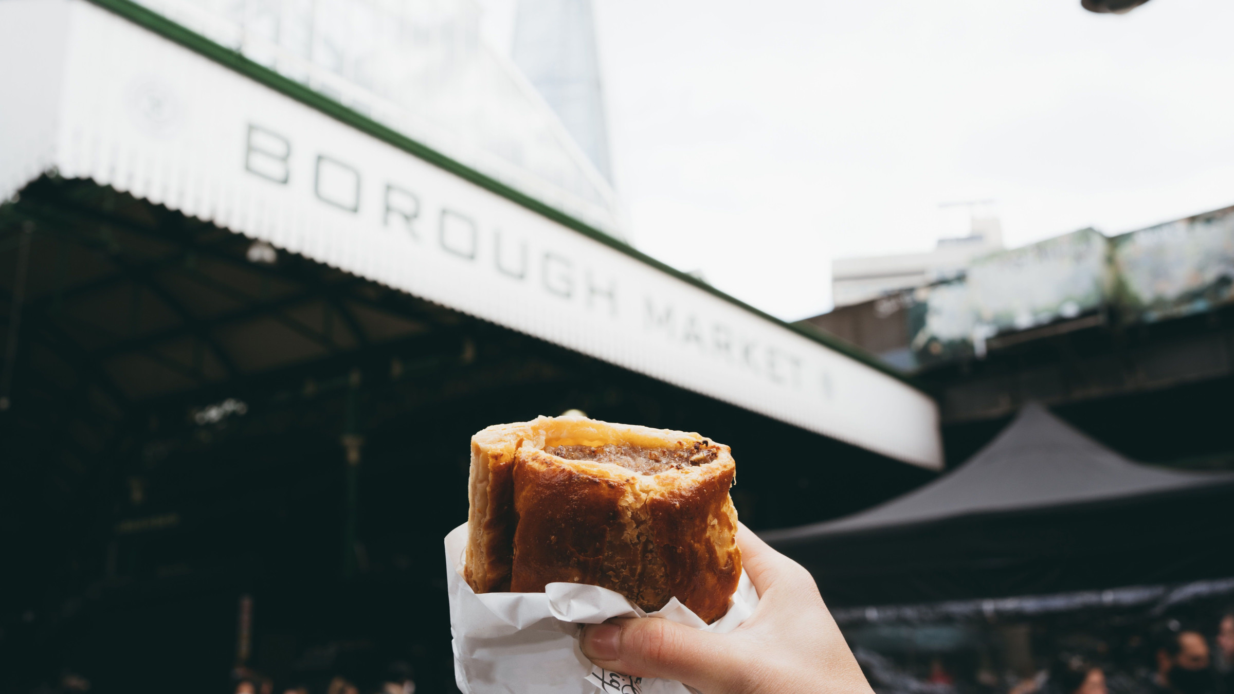 A hand holding a sausage roll in front of the entrance of the Borough Market in London. 
