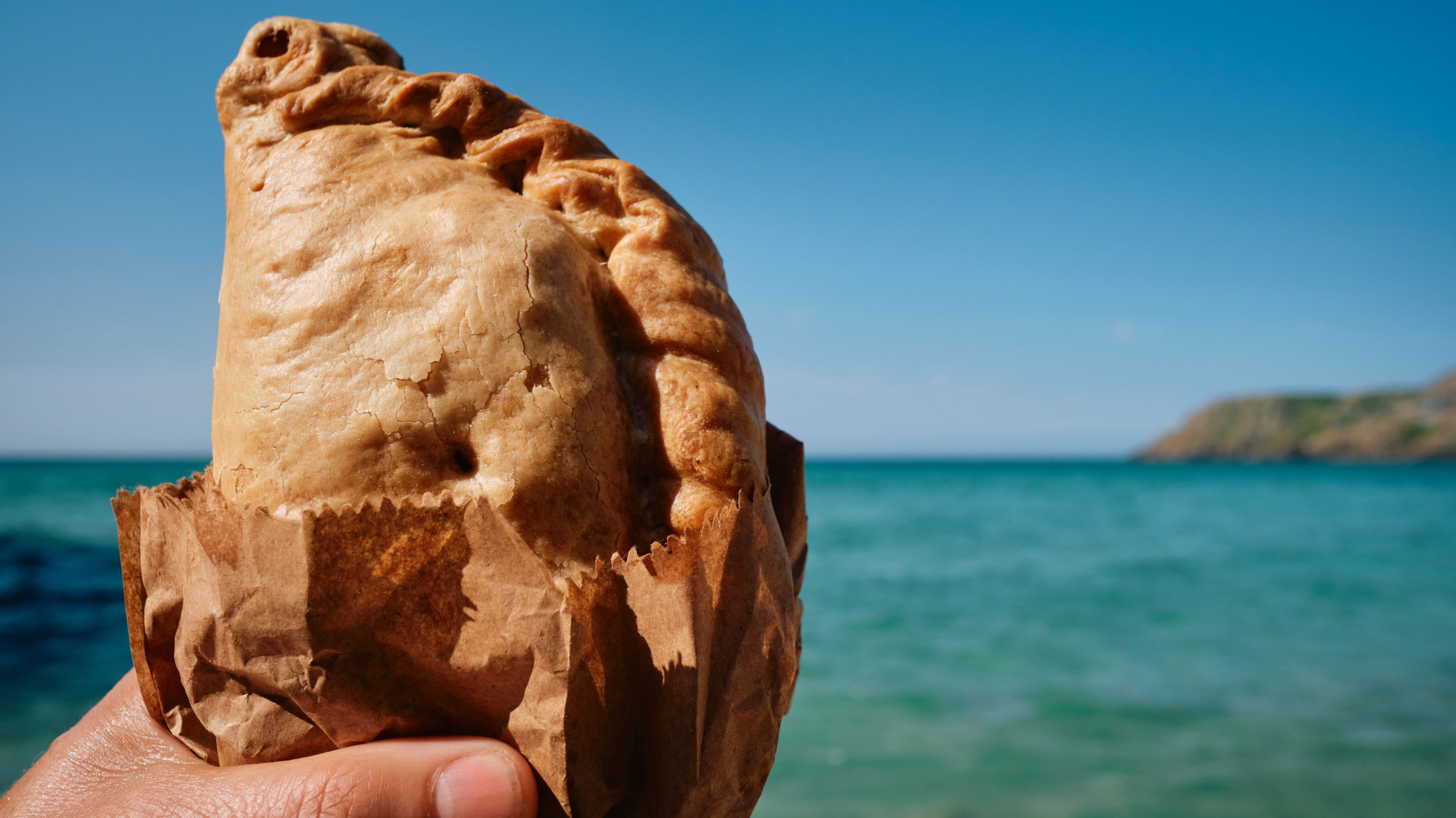 A hand holding a Cornish pasty with the sea in the background. 