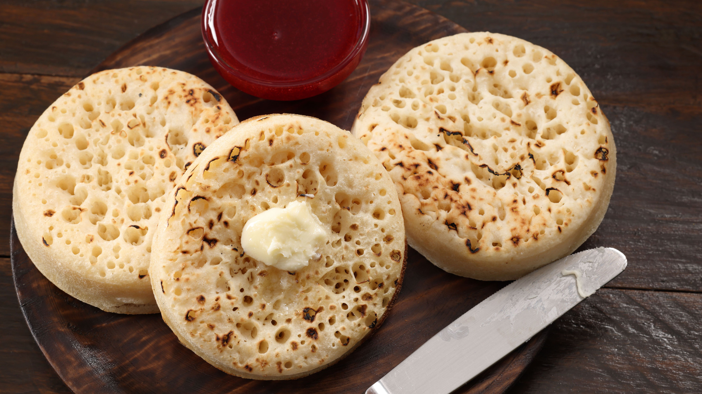Three crumpets. There's butter on top of one of them and a small bowl of jam next to them. 