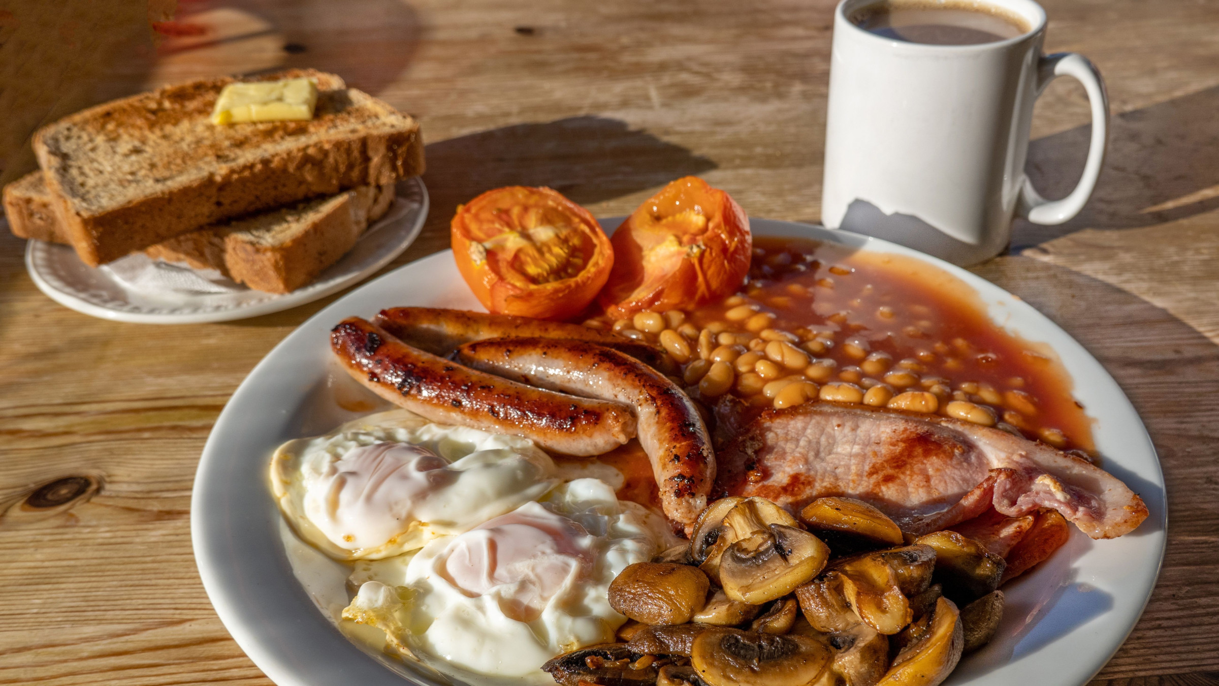 A close-up of a full English breakfast with fried eggs, sausages, bacon, tomatoes, beans, and mushrooms. There's also a plate with toasted bread and butter and a cup of coffee. 