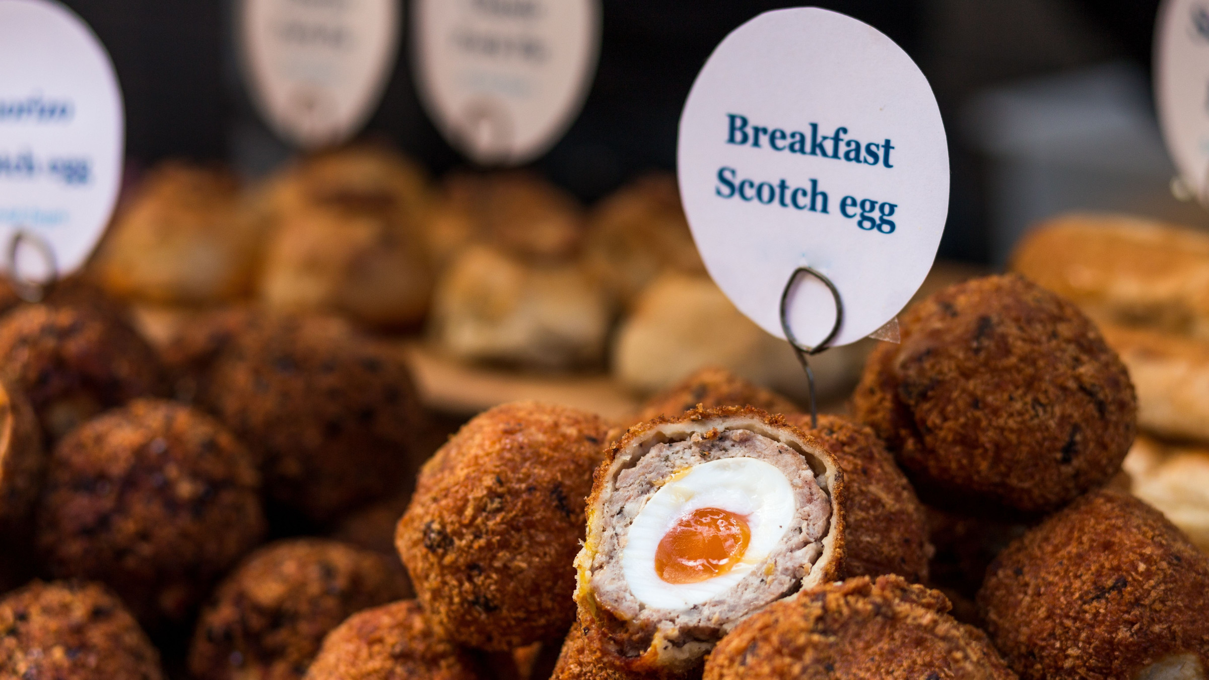 A pile of Scotch eggs with a small paper on top reading: Breakfast Scotch egg. 