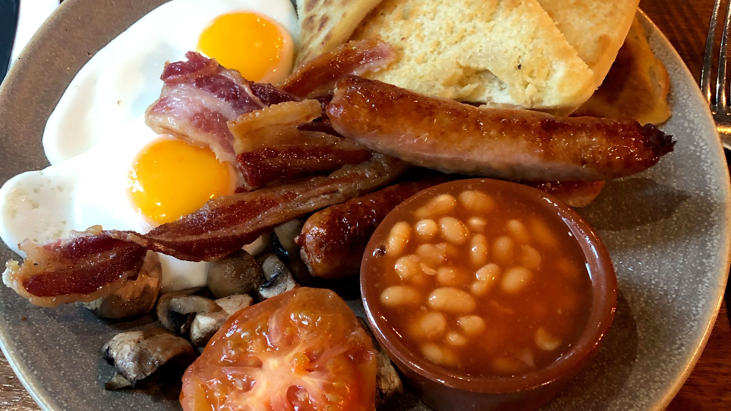 A close-up of an Ulster fry with toasted bread, two fried eggs, sausages, bacon, mushrooms, tomatoes, and beans. 