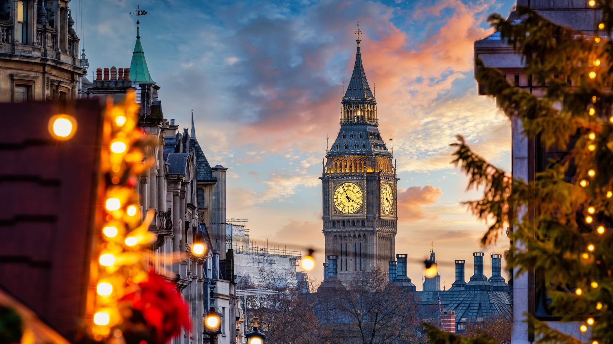 A beautiful sunset view of the Big Ben Clocktower in London, England, with the fairy lights from the Trafalgar Square Christmas market in the foreground.