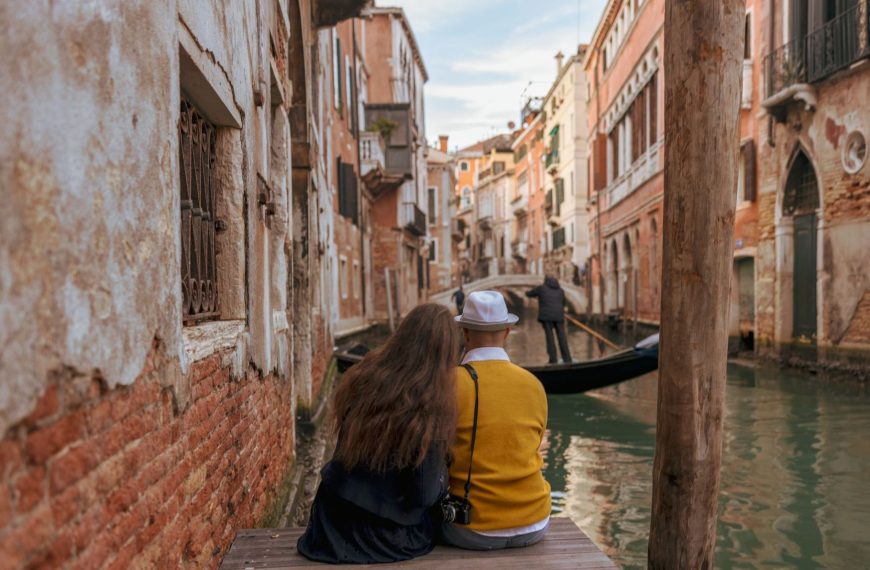 A couple with their backs turned to the camera are enjoying the view of a narrow canal, the city's traditional architecture, and a gondola in Venice.